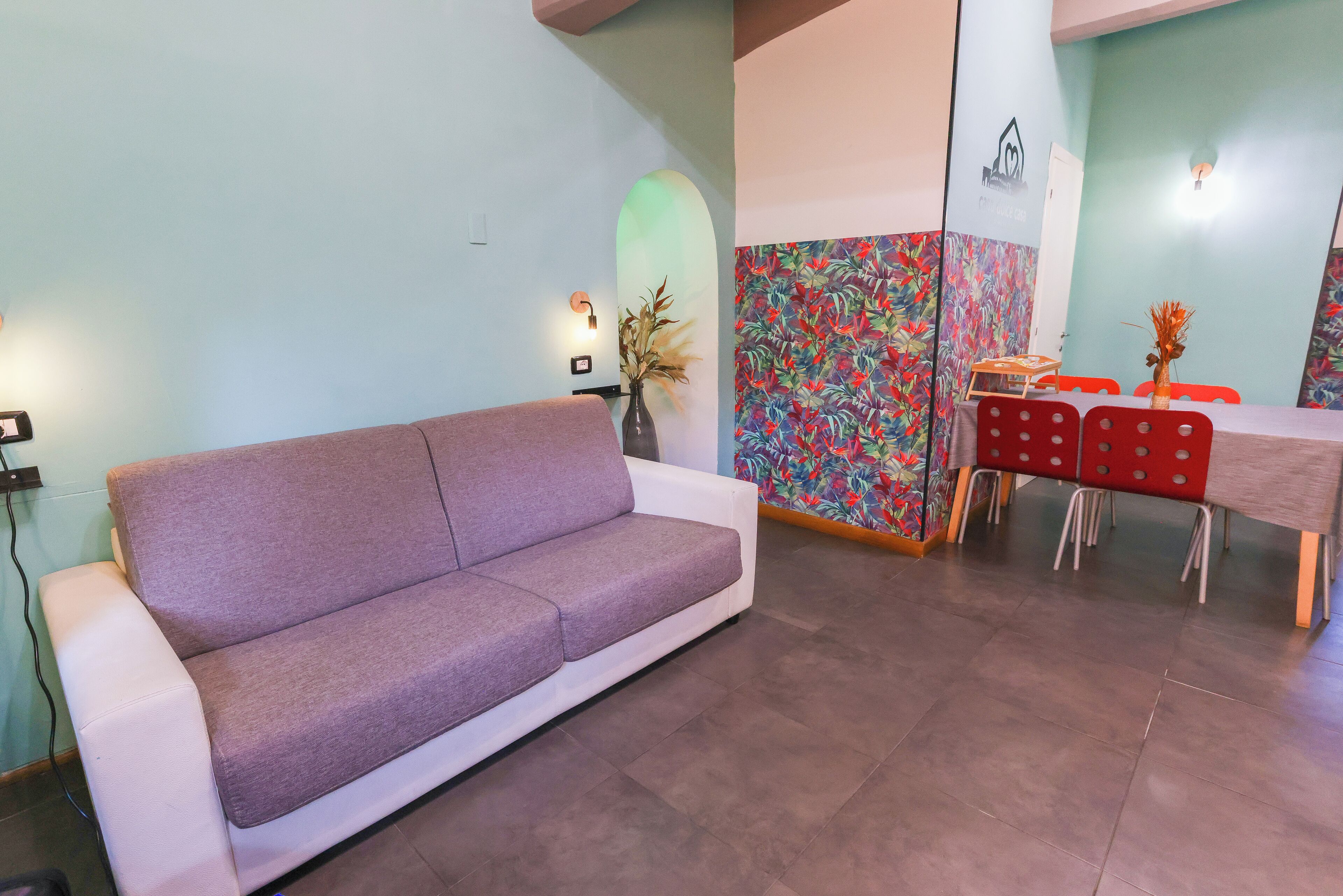 Ein modernes Wohnzimmer mit einem violetten Sofa, einem Esstisch mit roten Stühlen und einer farbenfrohen Wand mit Blumenmuster.