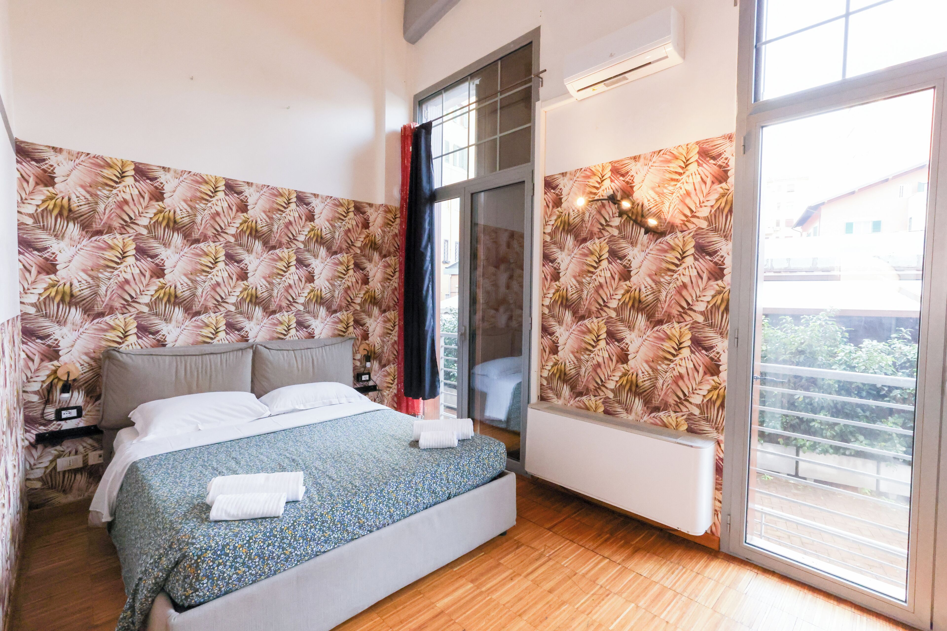 A bedroom with a bed, a nightstand, a window with a view, and a wall with a leaf pattern.