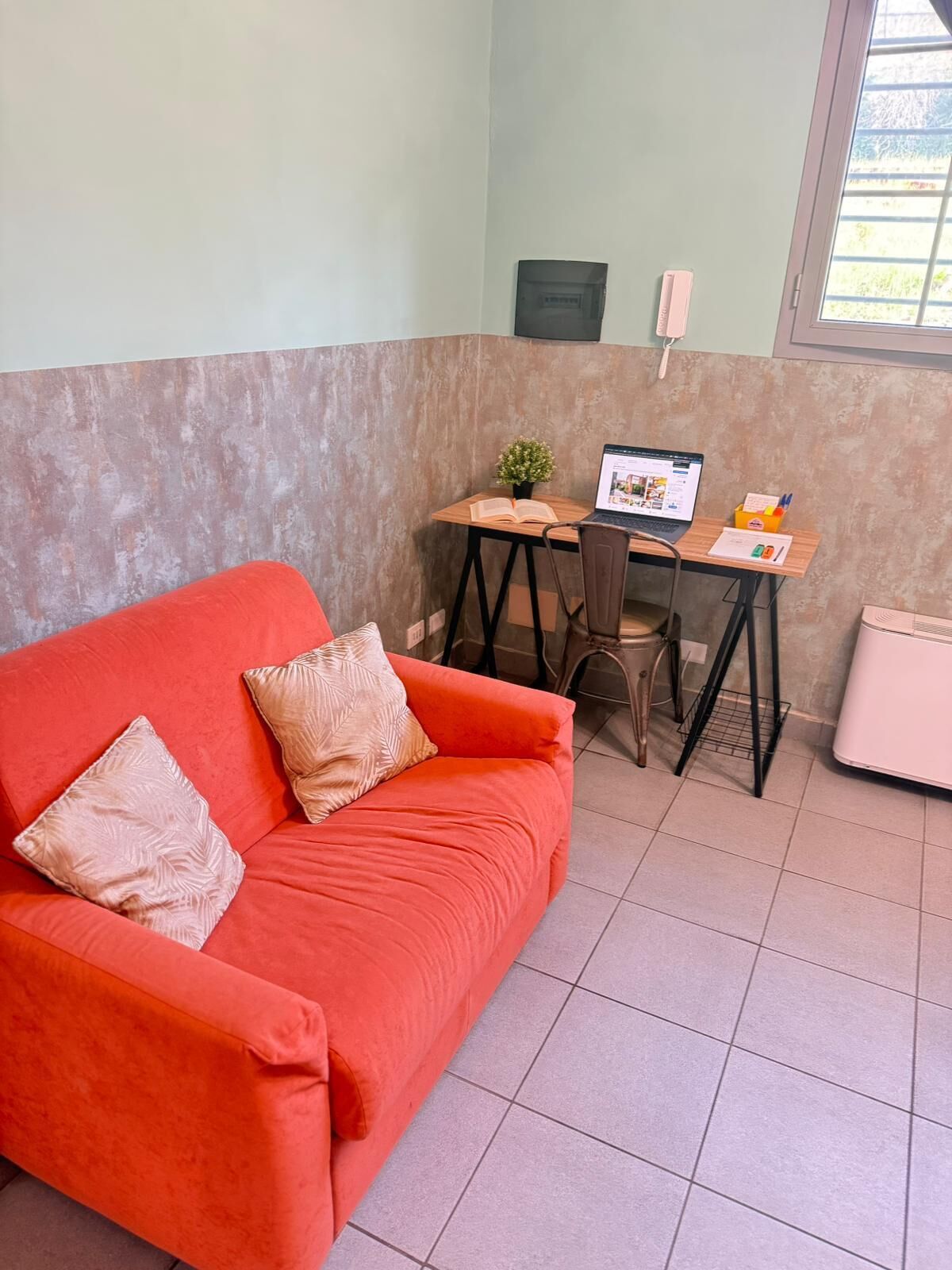 Une pièce avec un canapé rouge, un bureau en bois sur lequel se trouve un ordinateur portable, une petite plante en pot et un radiateur blanc.