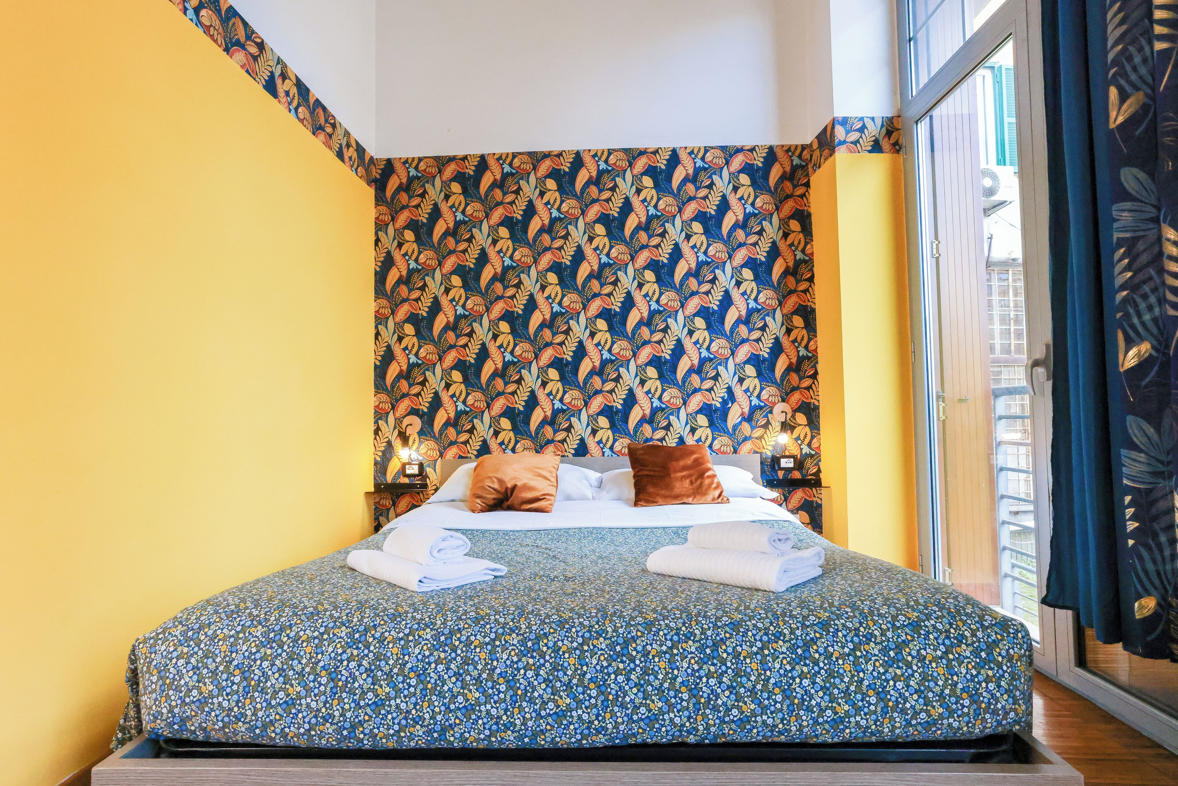 Ett hotellrum med ett blommigt sängöverkast, en gul accentvägg och utsikt genom ett fönster med gardiner.