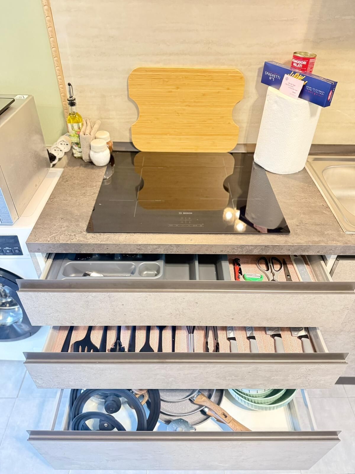 Una cocina con estufa empotrada, tabla de madera para cortar y diversos utensilios de cocina guardados en cajones.
