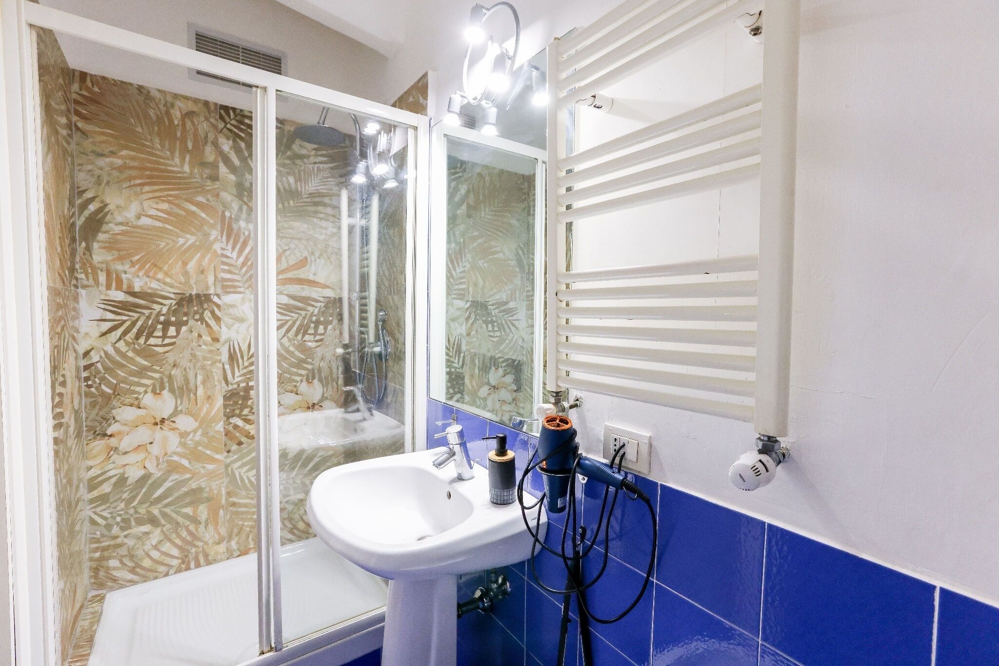 Ein Badezimmer mit einem weißen Waschbecken, blautonierten Fliesen, einer Duschkabine aus Glas und einem Spiegel mit weißem Handtuchhalter.