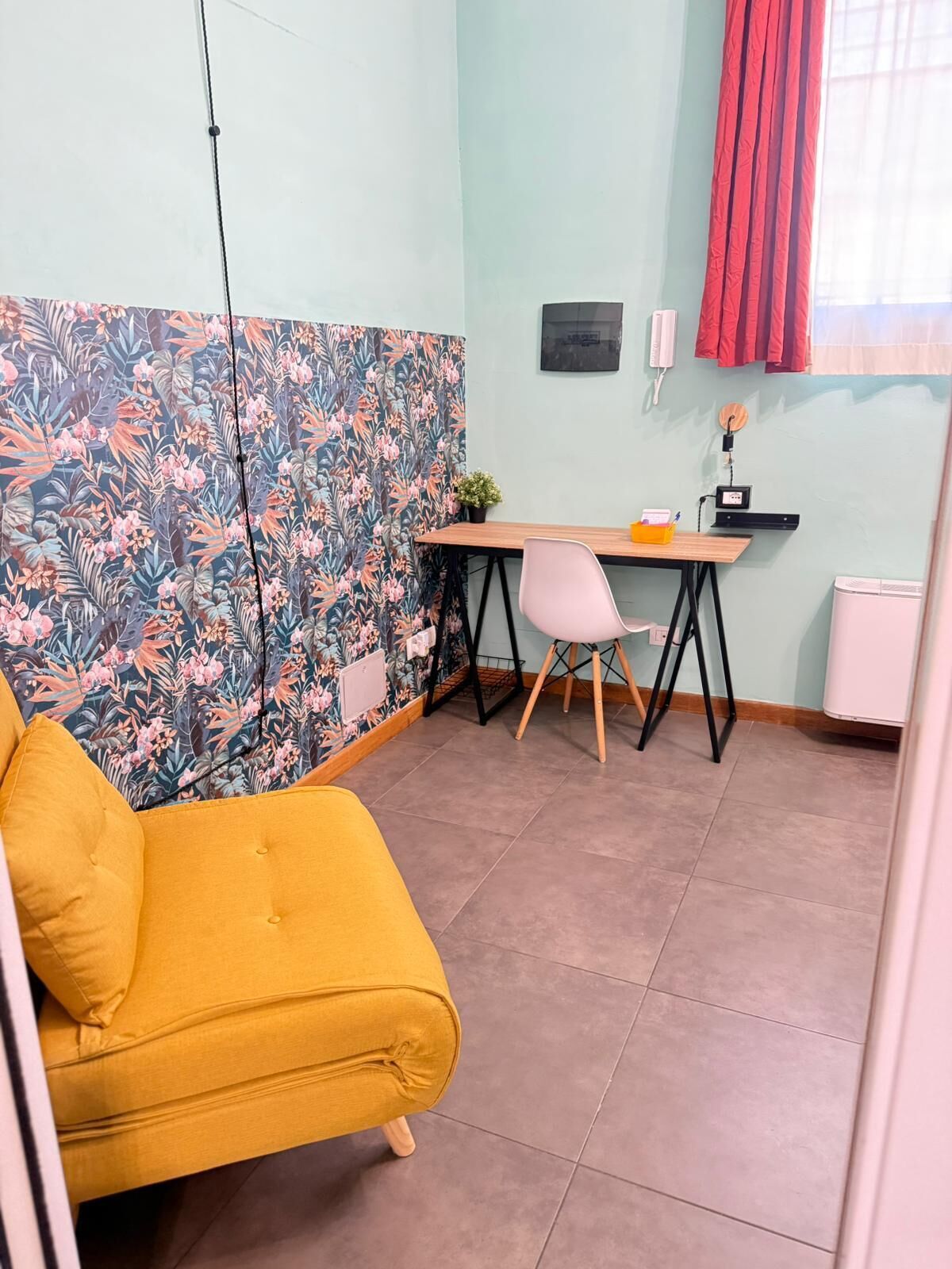 Une pièce avec un canapé jaune, un bureau avec une chaise, une petite plante et un rideau rouge.
