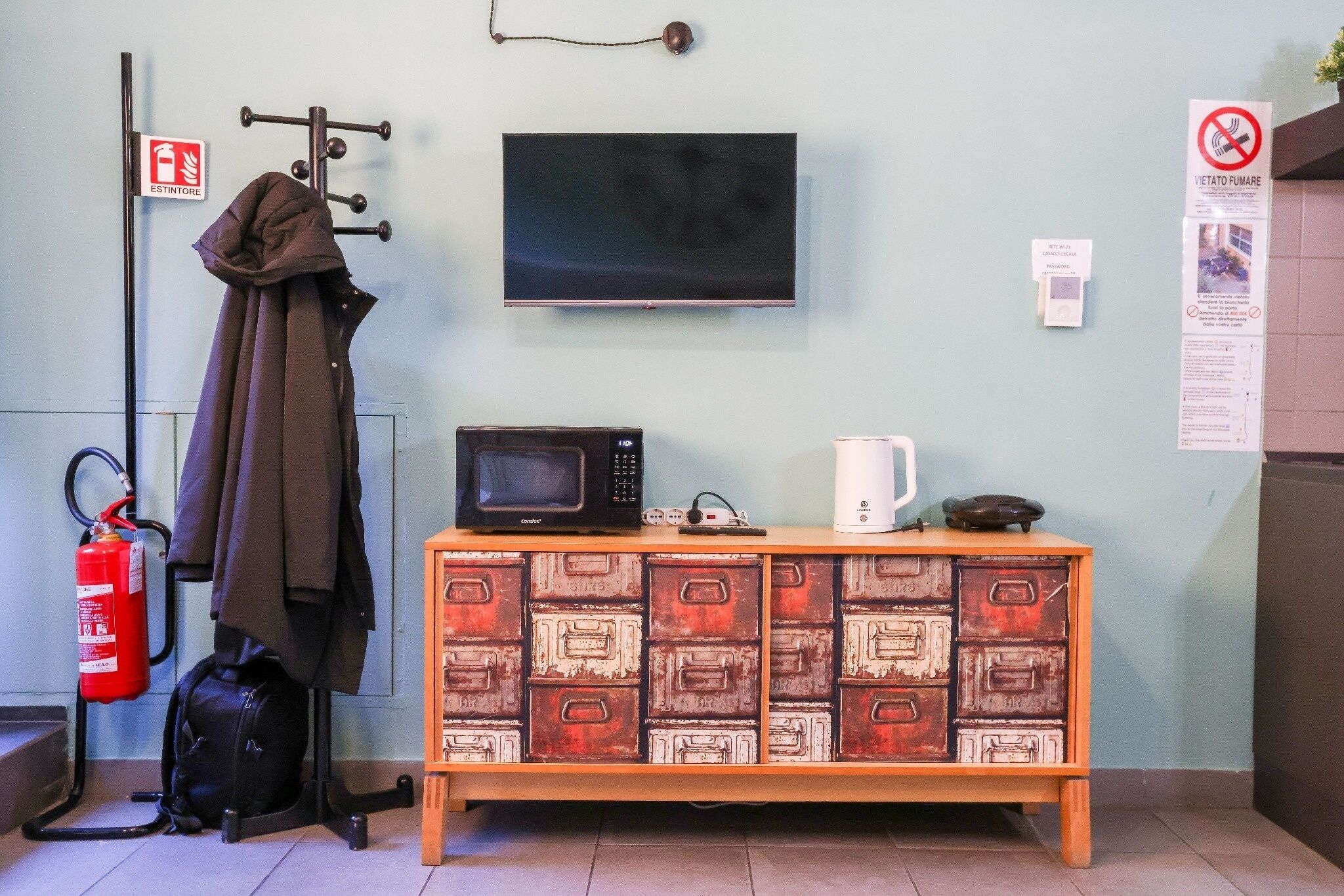 A room with a television mounted on the wall, a microwave on a wooden cabinet, a white mug, and a fire extinguisher on the floor.