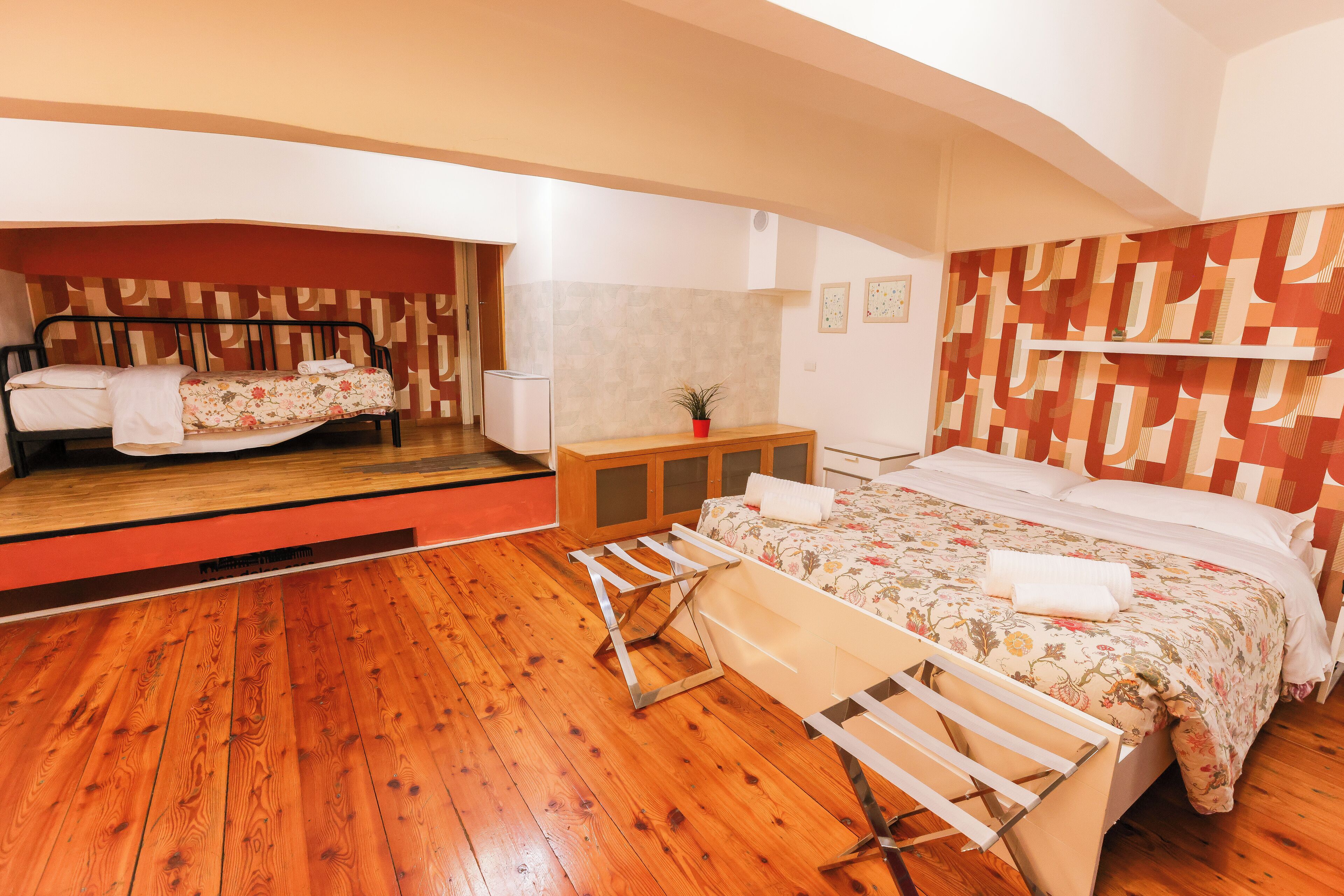 A bedroom with a wooden floor, a bed with floral bedding, a small bedside table with a plant, and a mirror above the bed.