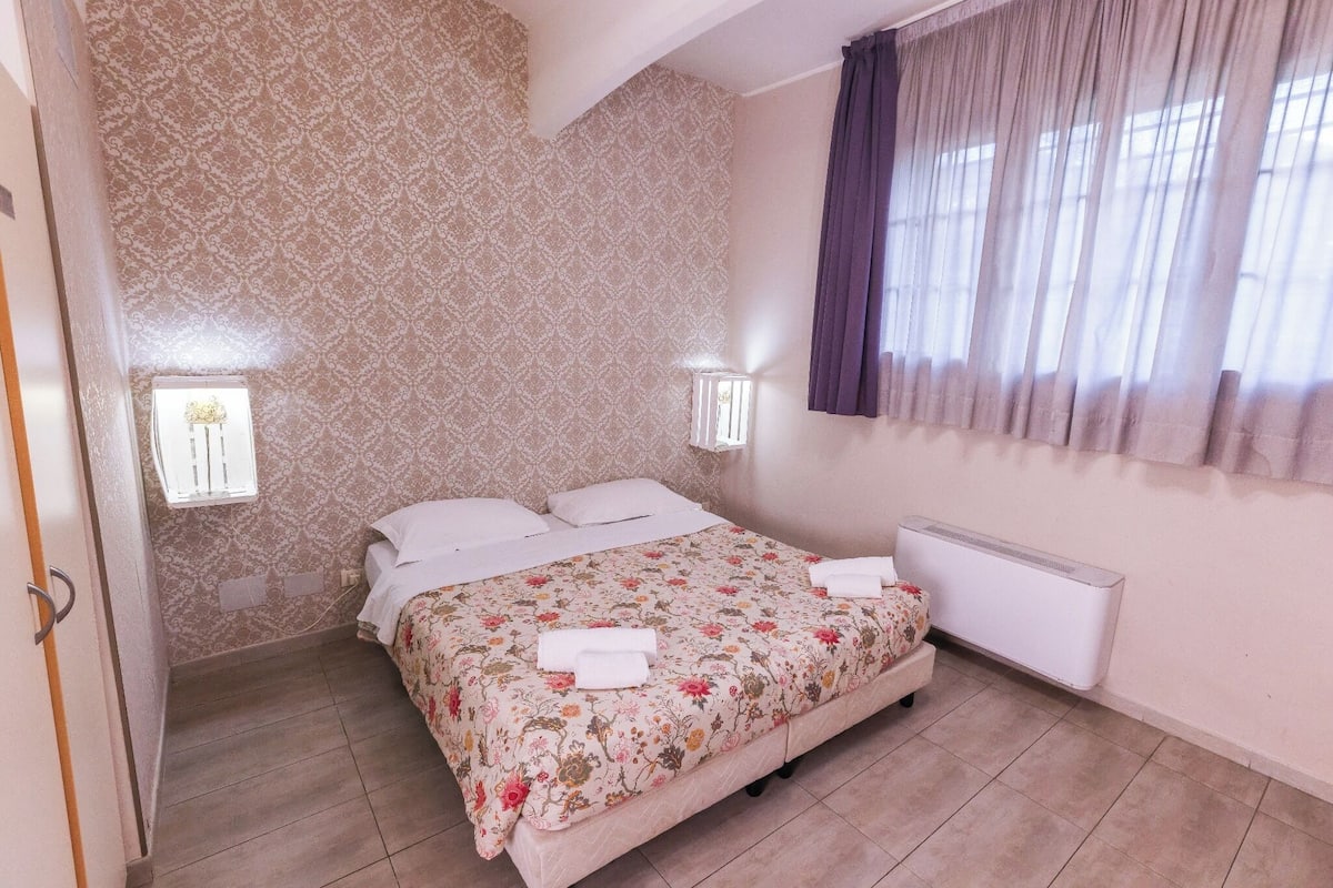A bedroom with a floral bedspread, two bedside lamps, a window with curtains, and a radiator.