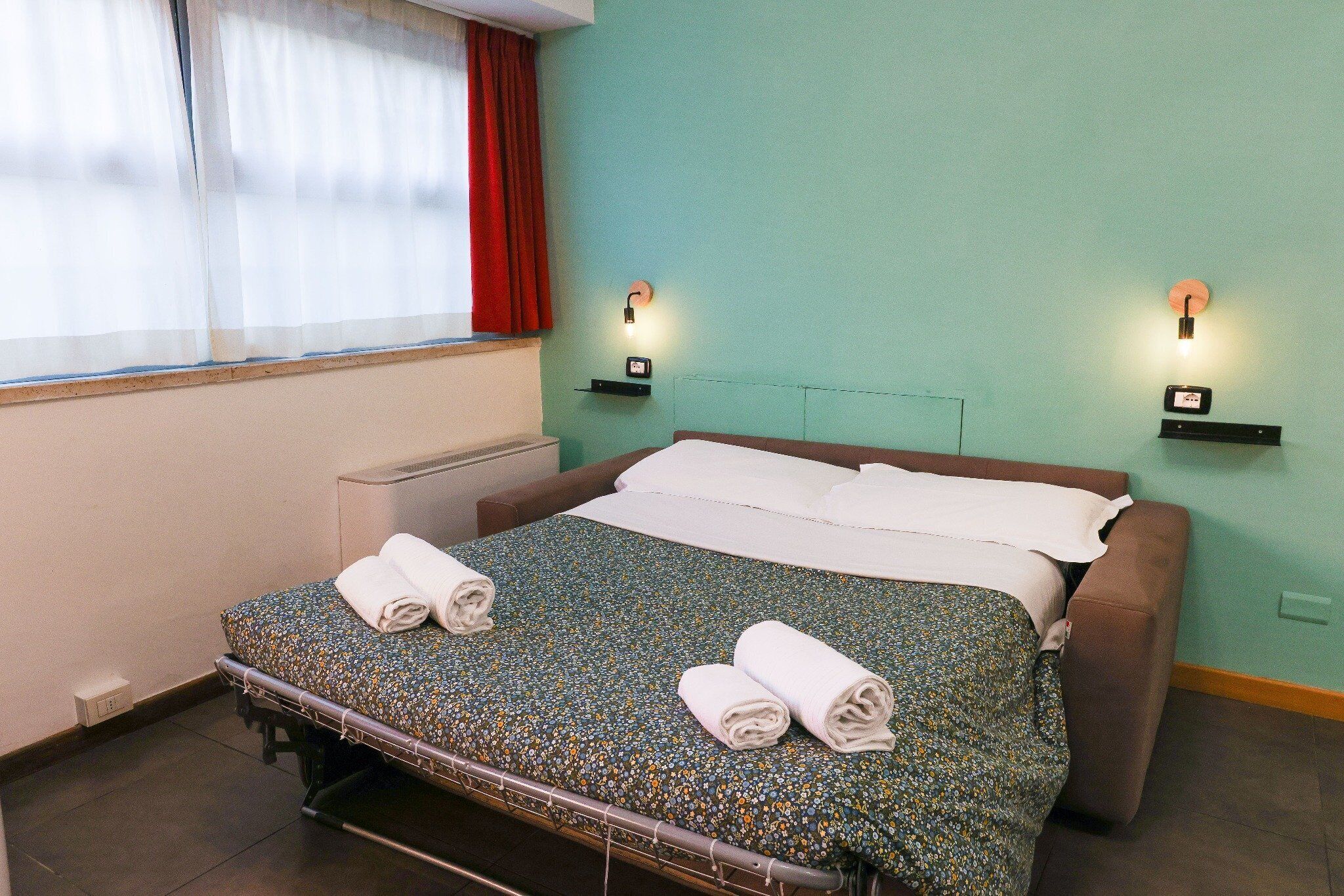 Een enkel bed met twee opgerolde handdoeken, een nachtkastje met een lamp en een raam met gordijnen.