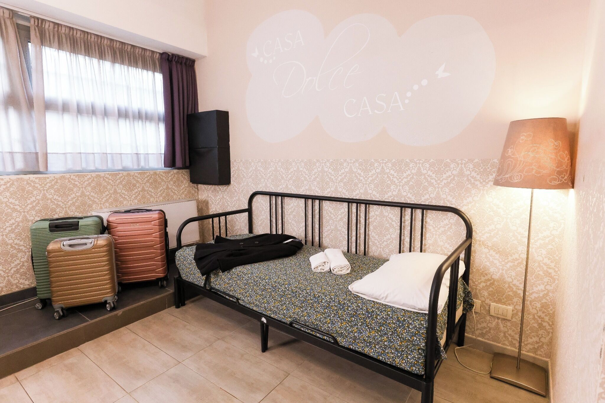 Una cama individual con colcha estampada, una lámpara de noche y una pared con una frase decorativa. Hay maletas y un altavoz en la pared.