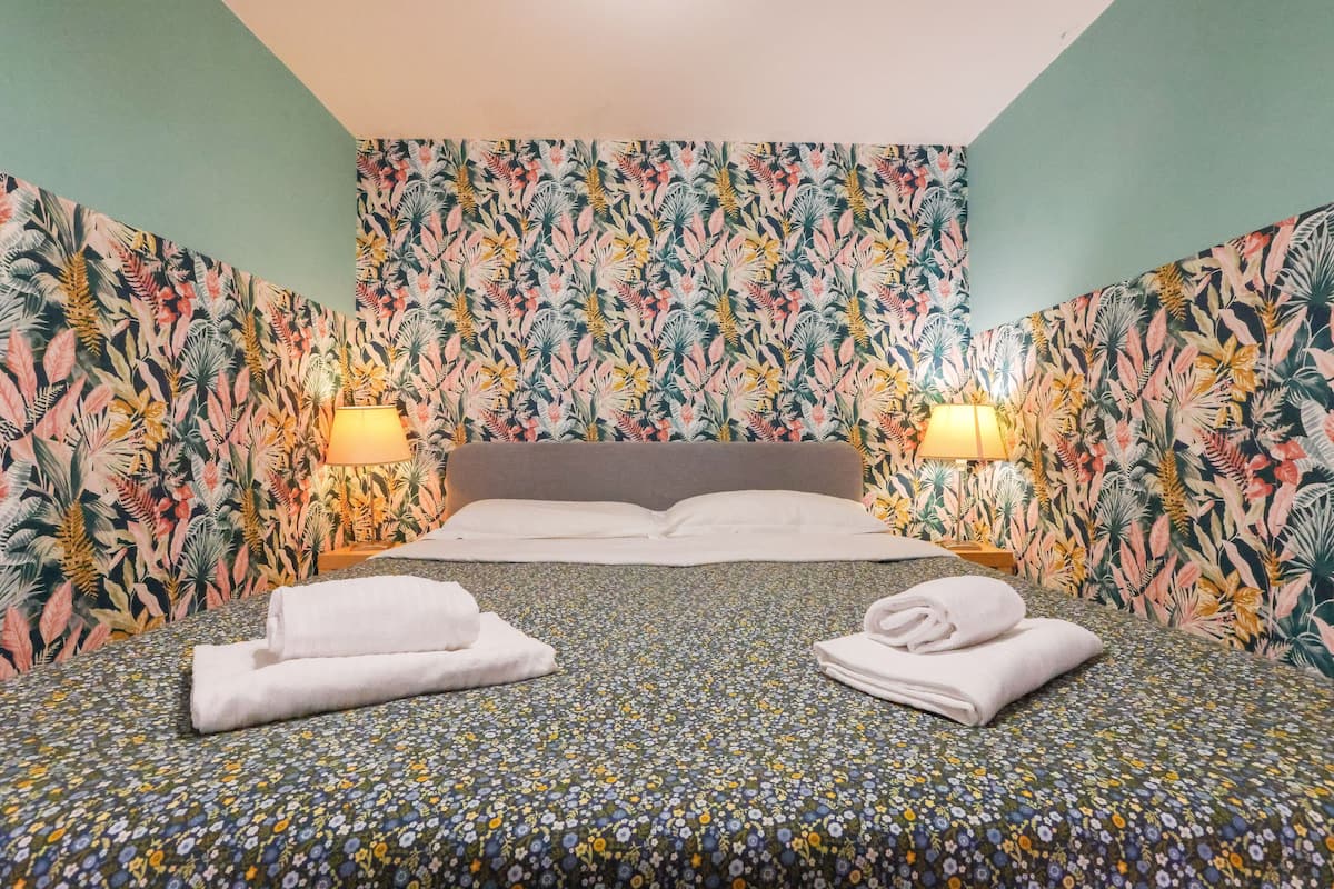 A hotel room with a floral-patterned wallpaper, a bed with a floral bedspread, two white towels on the bed, and two wall-mounted lamps.
