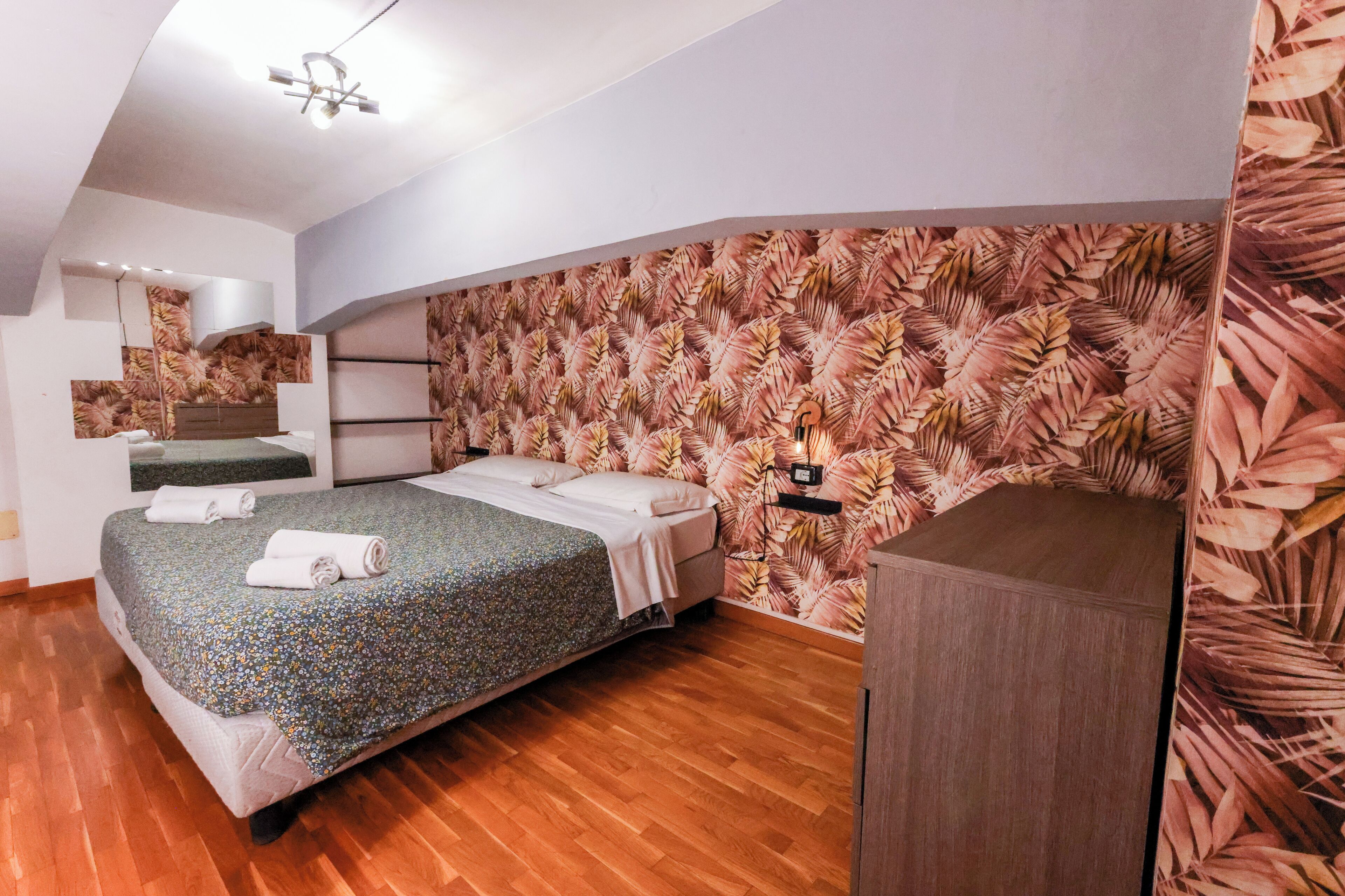 A bedroom with a bed, wooden flooring, a wall with a tropical leaf pattern, a bedside table, and a small cabinet.