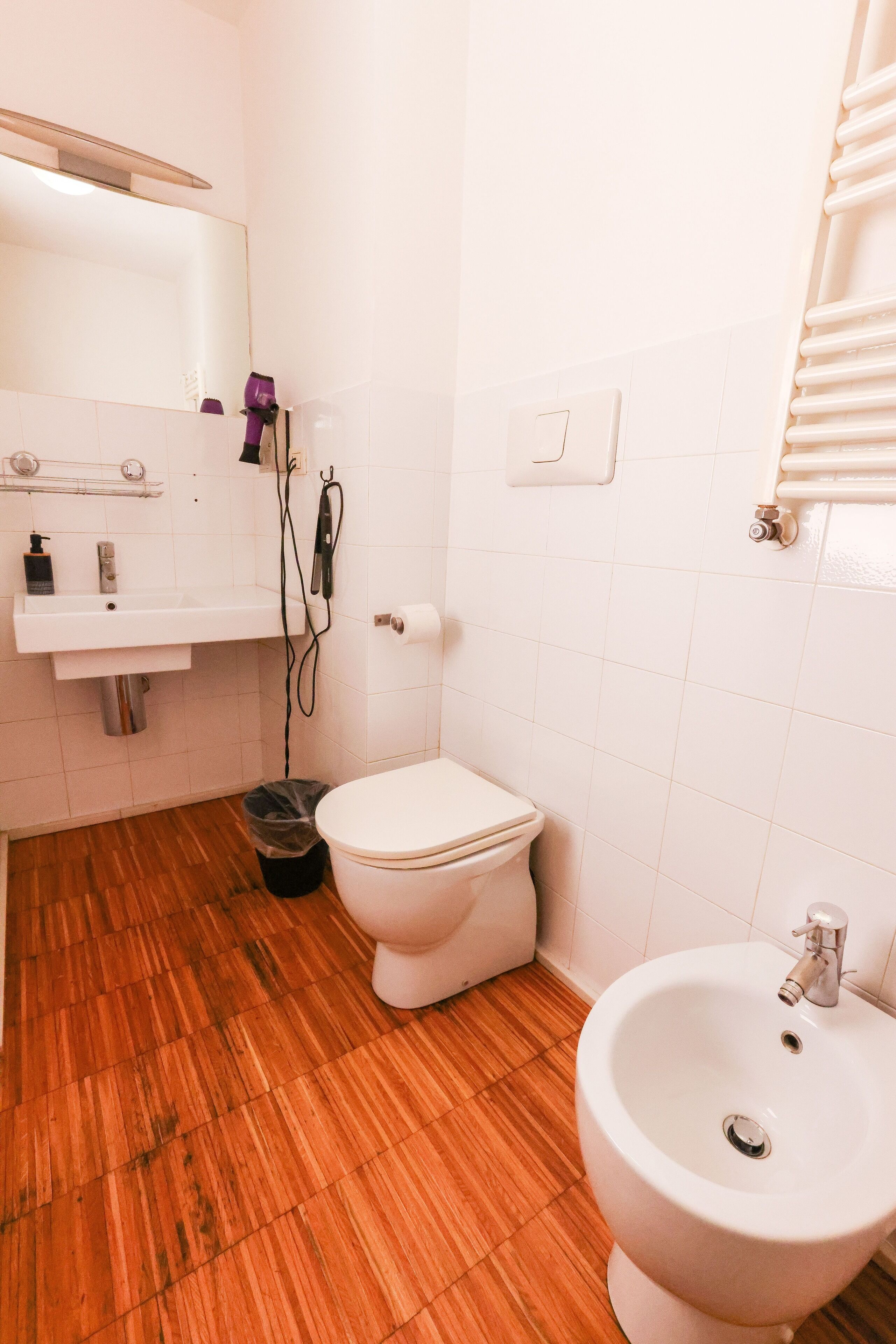 Uma casa de banho com chão de madeira, azulejos brancos, uma pia, uma sanita e um bidé.