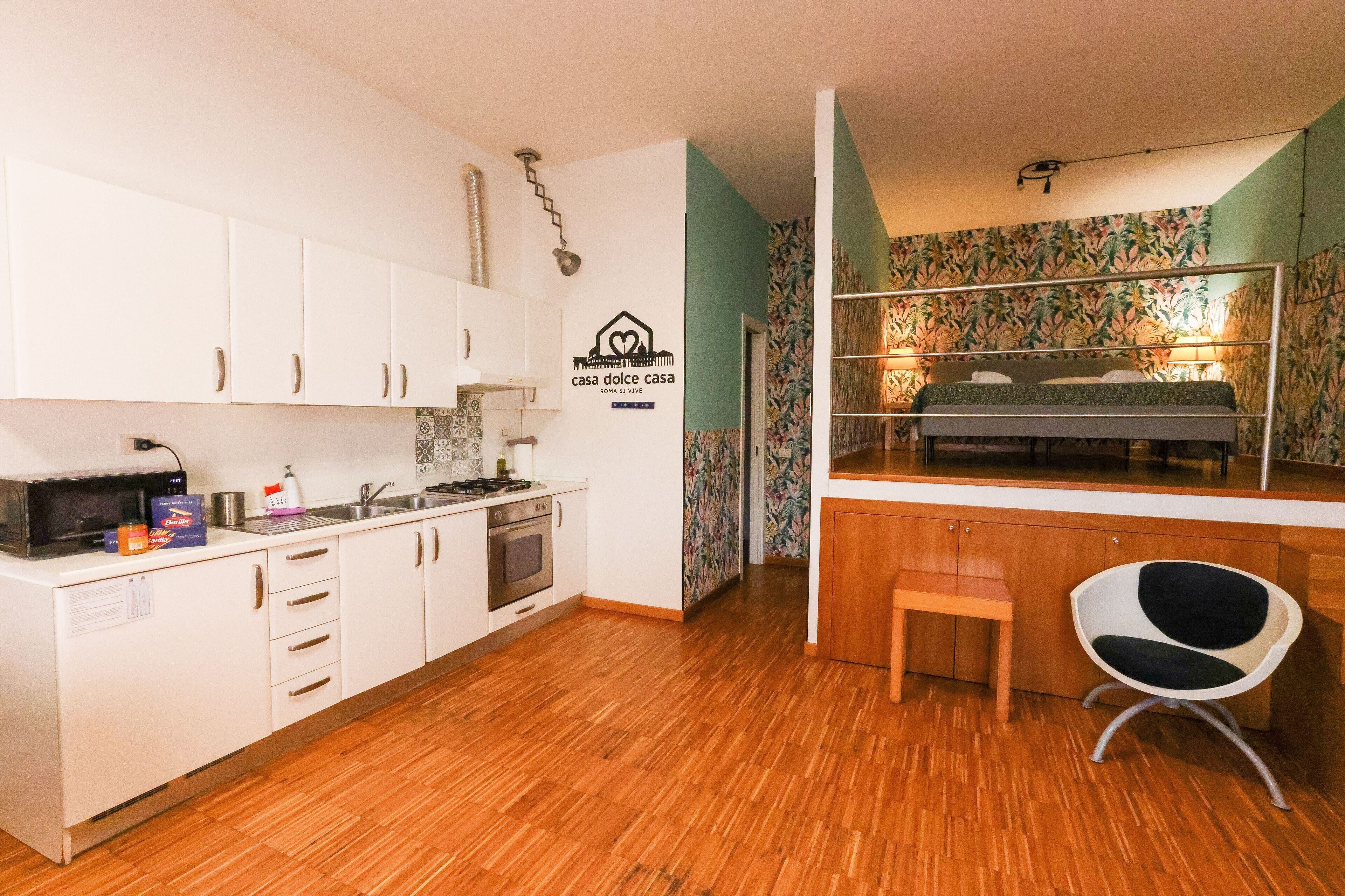Una cucina compatta con mobili bianchi, un forno a microonde e un piano cottura. Si trova un piccolo tavolo di legno e una sedia con seduta nera.