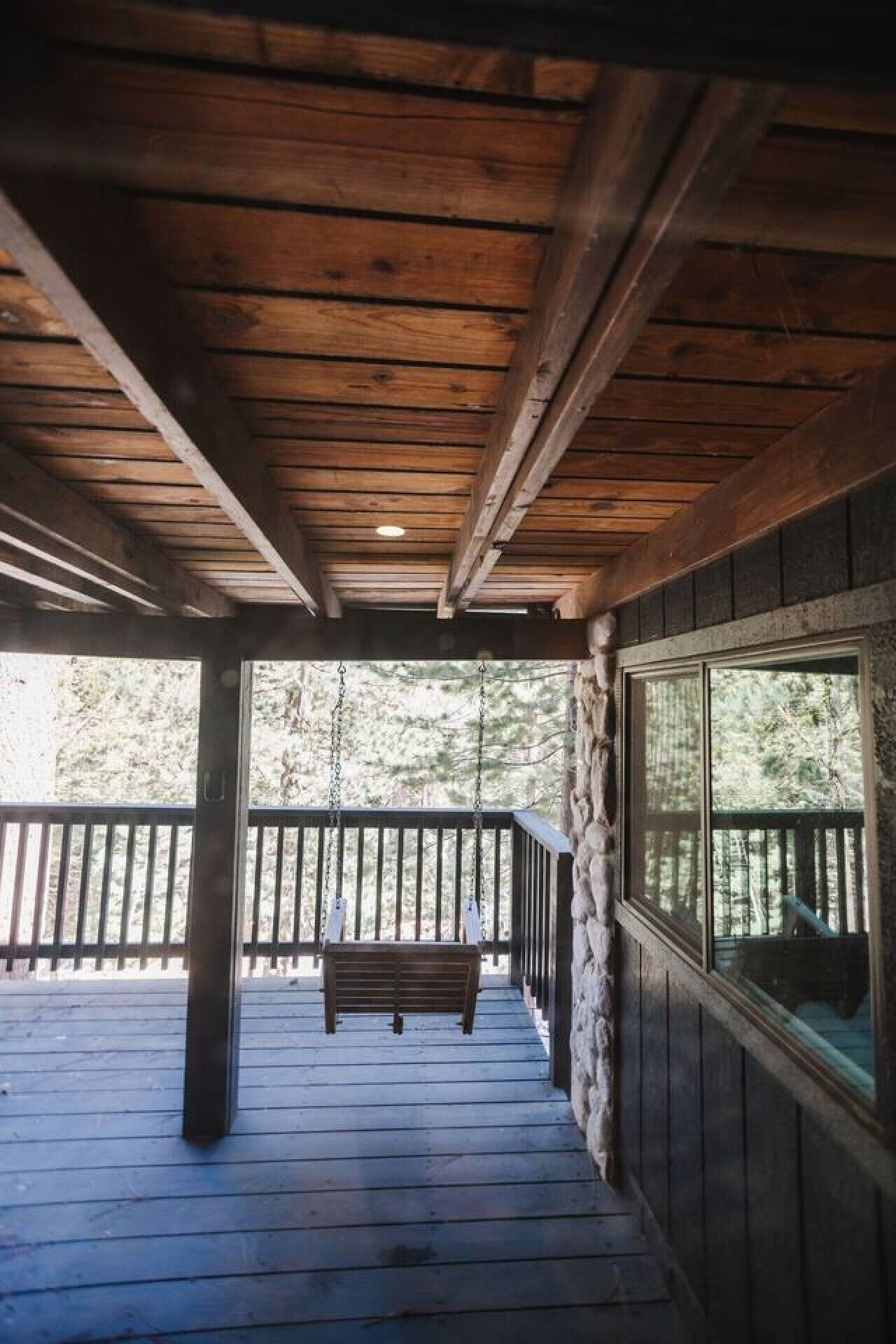 Una terraza de madera con un banco, un muro de piedra y una ventana con vista a un bosque.