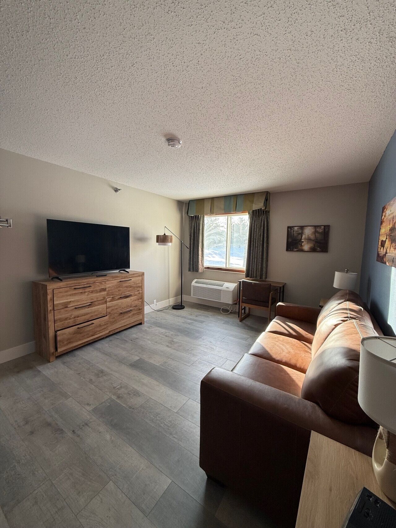 A living room with a wooden TV stand, a leather sofa, a floor lamp, and a window with curtains.