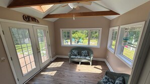 Una habitación con piso de madera, ventilador de techo, ventana con vista a un jardín y una silla de mimbre.