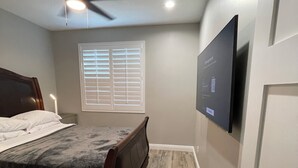 Un dormitorio con cama, ventilador de techo, ventana con persianas y televisor de pantalla plana montado en la pared.
