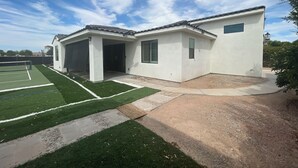 Una casa de una planta con un porche cubierto, cancha de tenis y camino de entrada de tierra.