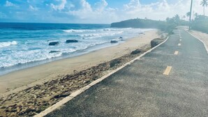 Camí costaner amb una platja d'un costat i un carrer amb palmeres i una creu a l'altre.