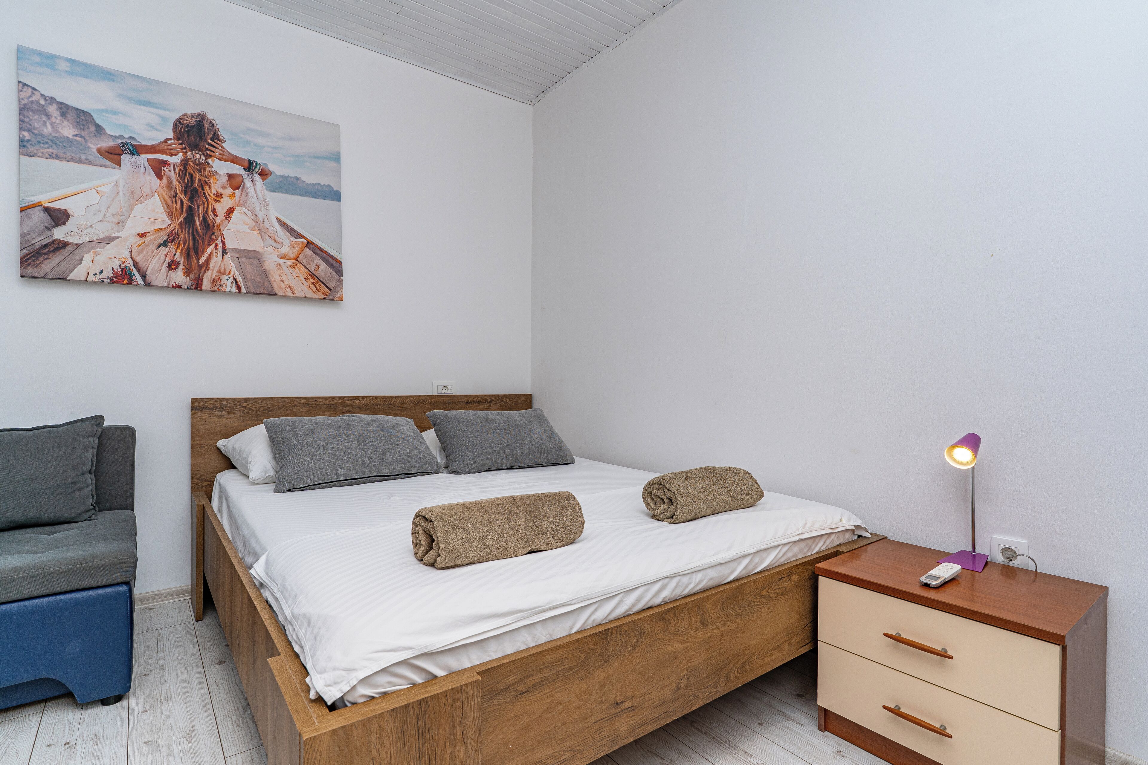 A bedroom with a wooden bed, a bedside table with a lamp, a small sofa, and a painting on the wall.