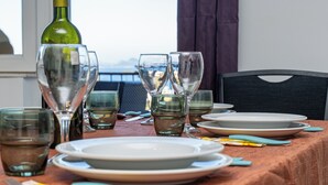Une table à manger dressée avec des assiettes, des verres et une bouteille de vin, avec vue sur la mer par une fenêtre.