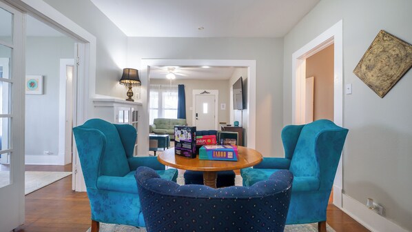 Un salon avec des fauteuils turquoise, une table ronde en bois et un pouf à motifs. On y trouve des boîtes sur la table, une lampe et un élément décoratif mural.