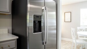 Un refrigerador de acero inoxidable con dispensador de agua, una encimera blanca y un comedor con sillas y mesa blancos.