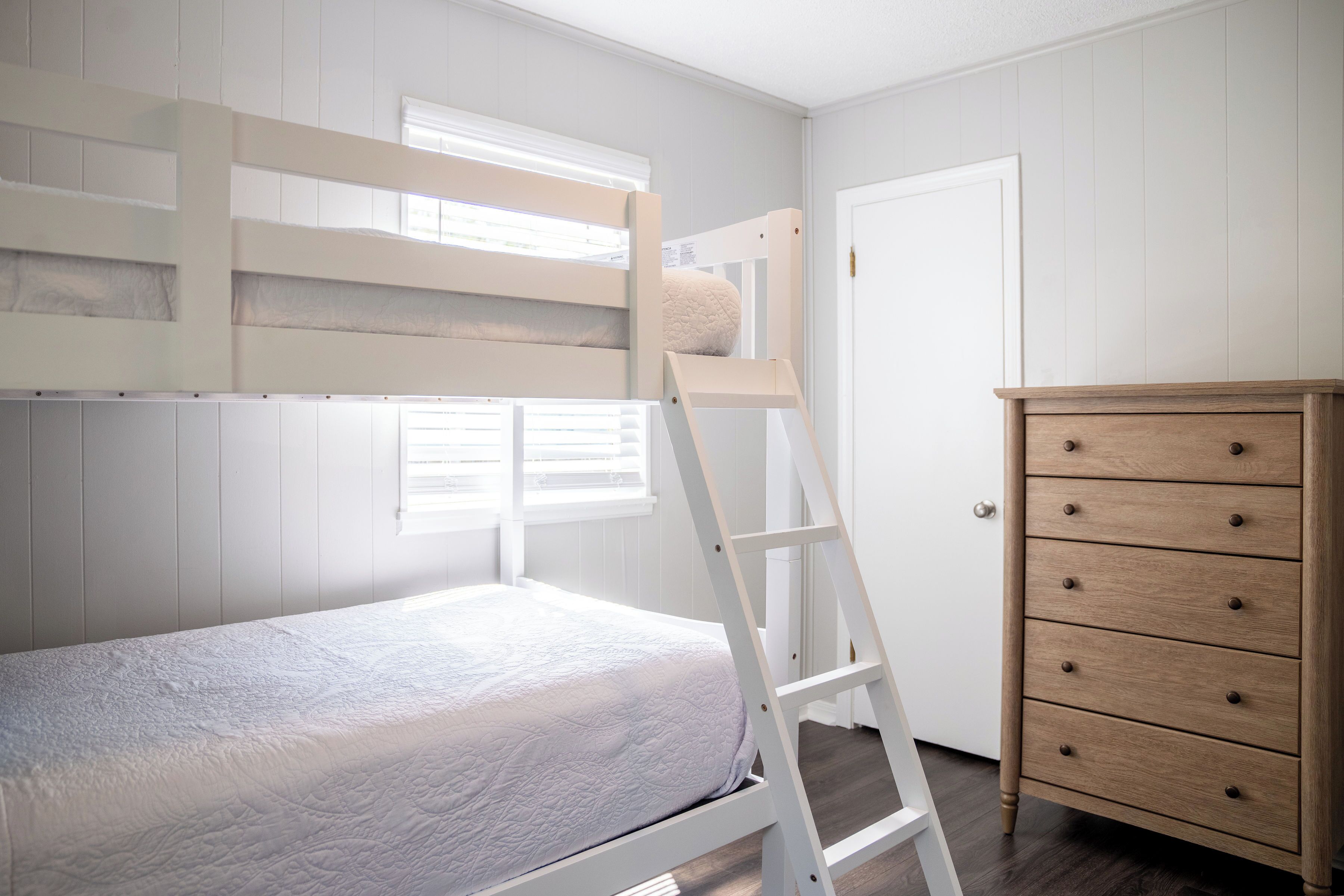 A bunk bed with a ladder, a wooden dresser, and a white door in a room with white walls and wooden flooring.