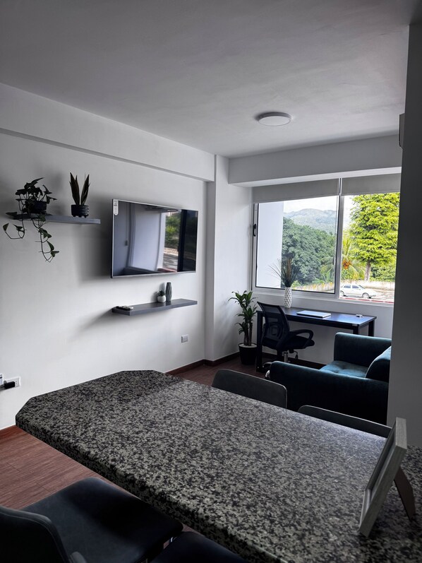 Una habitación moderna con televisión de pantalla plana, un escritorio con silla, un sofá y una ventana con vistas a zonas verdes.