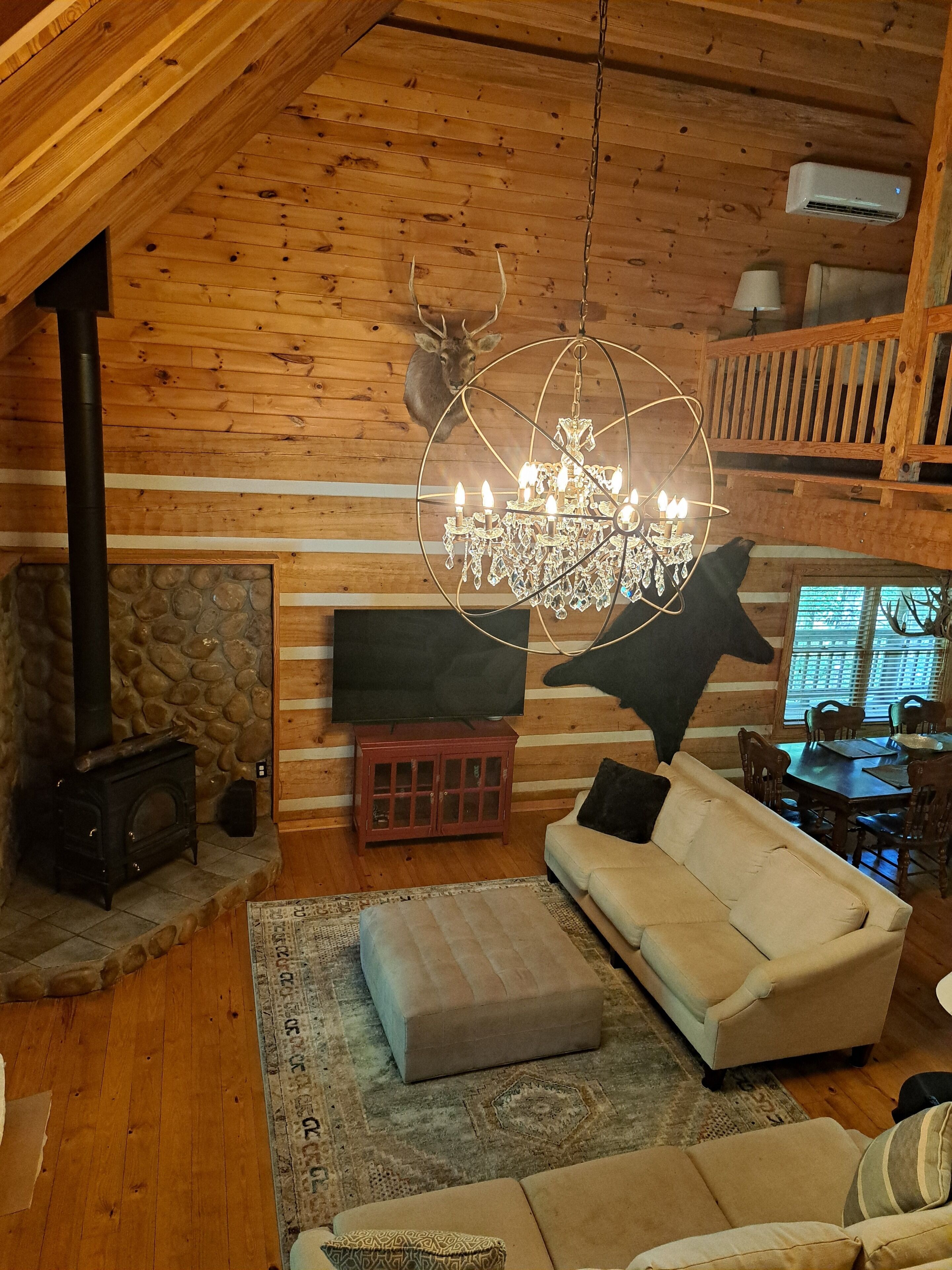 A cozy cabin interior with a fireplace, a chandelier, a mounted deer head, and a dining area.