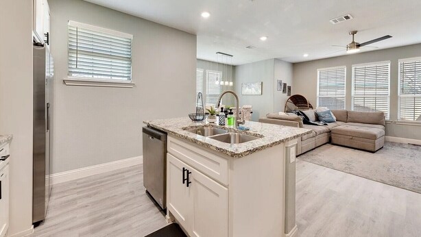 Una cocina moderna con lavavajillas incorporado, encimera de granito y una isla grande con fregadero. Hay una zona de comedor con sofá y ventilador de techo.