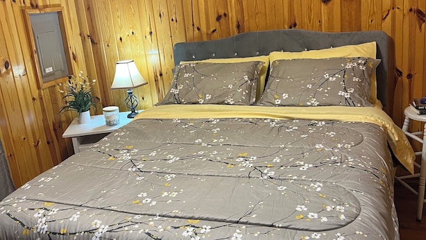 A bedroom with a wooden wall, a bed with floral bedding, a nightstand with a lamp, and a sign above the bed.
