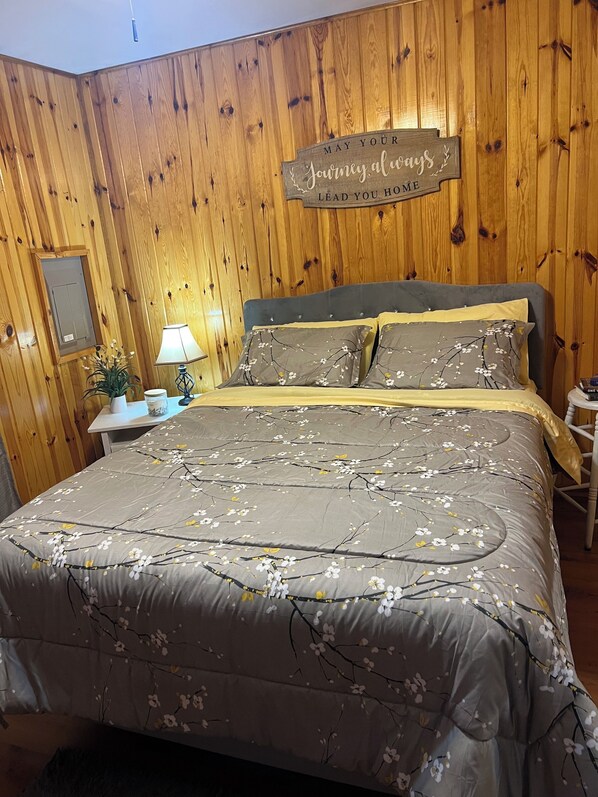 Une chambre avec un mur en bois, un lit recouvert d’une literie à motifs floraux, une table de chevet avec une lampe, et un panneau décoratif au-dessus du lit.