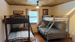 A room with two bunk beds, wooden flooring, a ceiling fan, a window with curtains, and a framed picture on the wall.