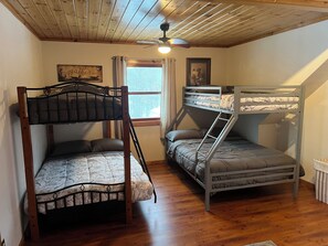 Une chambre avec deux lits superposés, un plancher en bois, un ventilateur de plafond, une fenêtre avec des rideaux et un tableau encadré au mur.