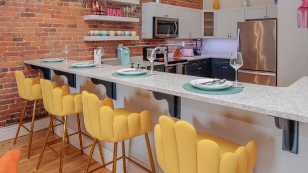 Una cocina con pared de ladrillo, armarios blancos, un refrigerador de acero inoxidable y taburetes amarillos.