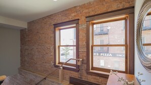 Una habitación con paredes de ladrillo a la vista, piso de madera y ventanas amplias que dan a un edificio de ladrillo.