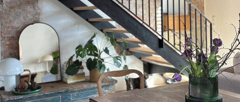 A wooden dining table with a vase of flowers, a staircase with a metal railing, and a brick wall in the background.
