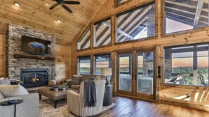 A spacious living room with a stone fireplace, large windows, and a ceiling fan. - Toccoa Vista Pool & Spa |Mineral Bluff, GA (Mineral Bluff)