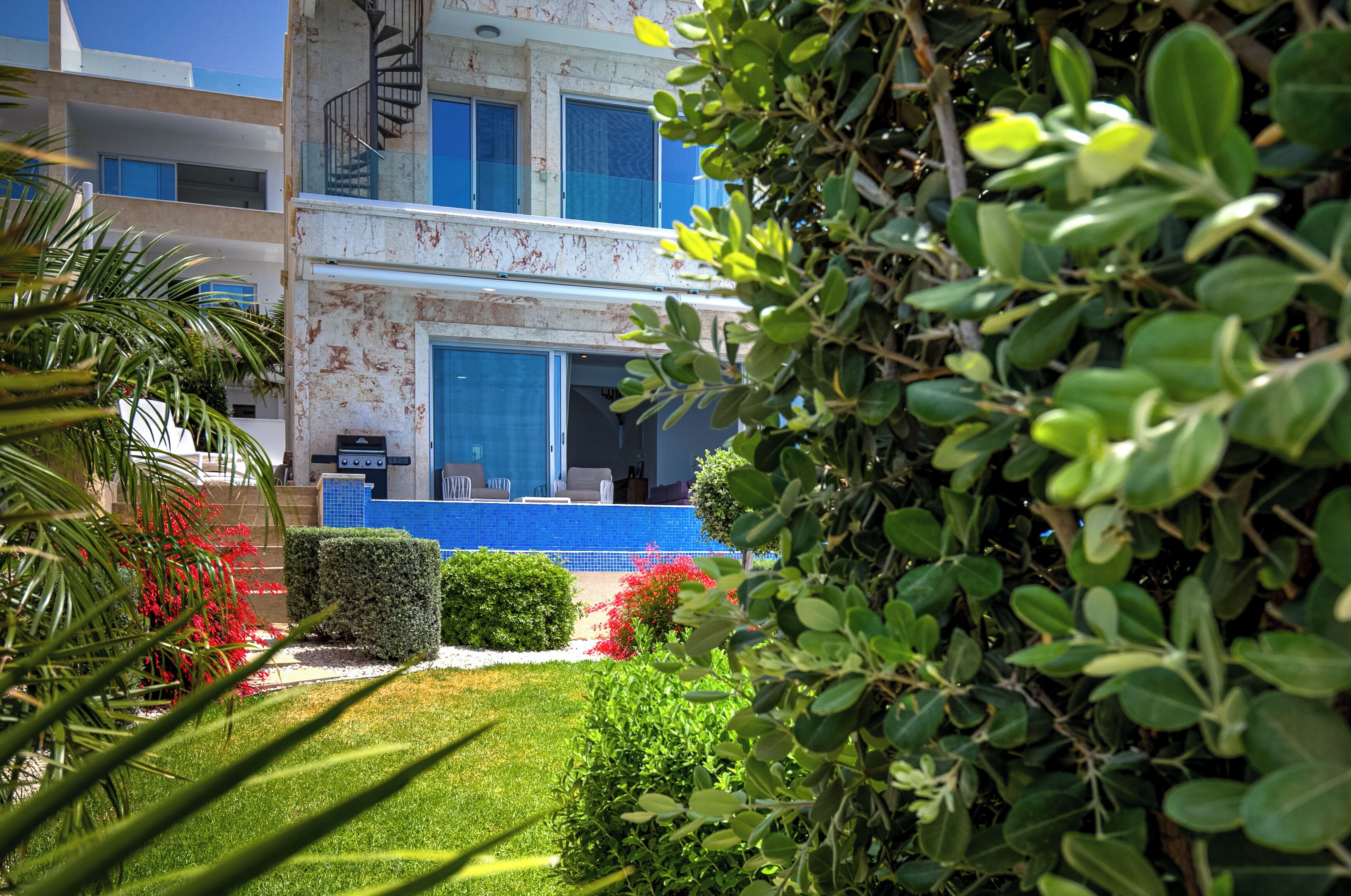 Un edificio de dos pisos con una piscina de azulejos azules, rodeado de exuberante vegetación y una escalera en espiral.