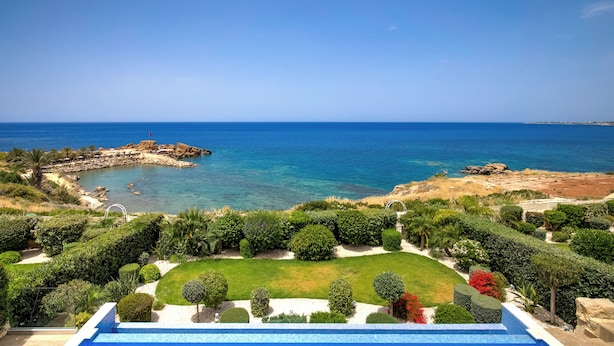 A landscaped garden with a pool, a rocky coastline, and a clear blue sea. - Olymp Luxury Villa D (Kissonerga)