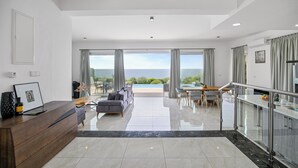 A modern living room with a large window, a sofa, a dining area, and a kitchen with a balcony. - Olymp Luxury Villa D (Kissonerga)