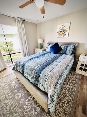 Un dormitorio con una cama grande, un ventilador de techo, una mesita de noche con lámpara y vistas al exterior a través de una puerta corrediza.