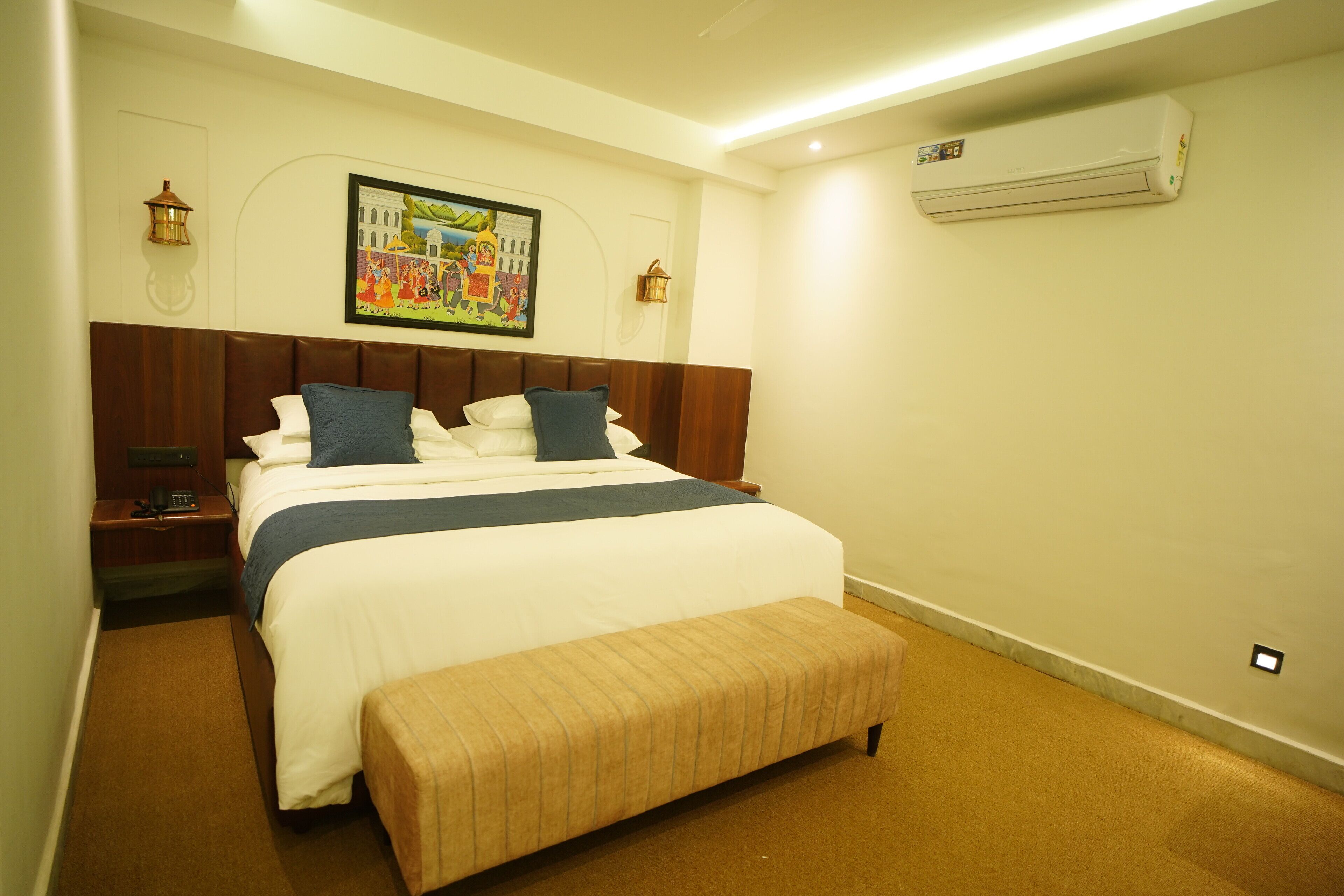 A hotel room with a large bed, a bench, a nightstand, a wall-mounted air conditioner, and a painting above the bed.