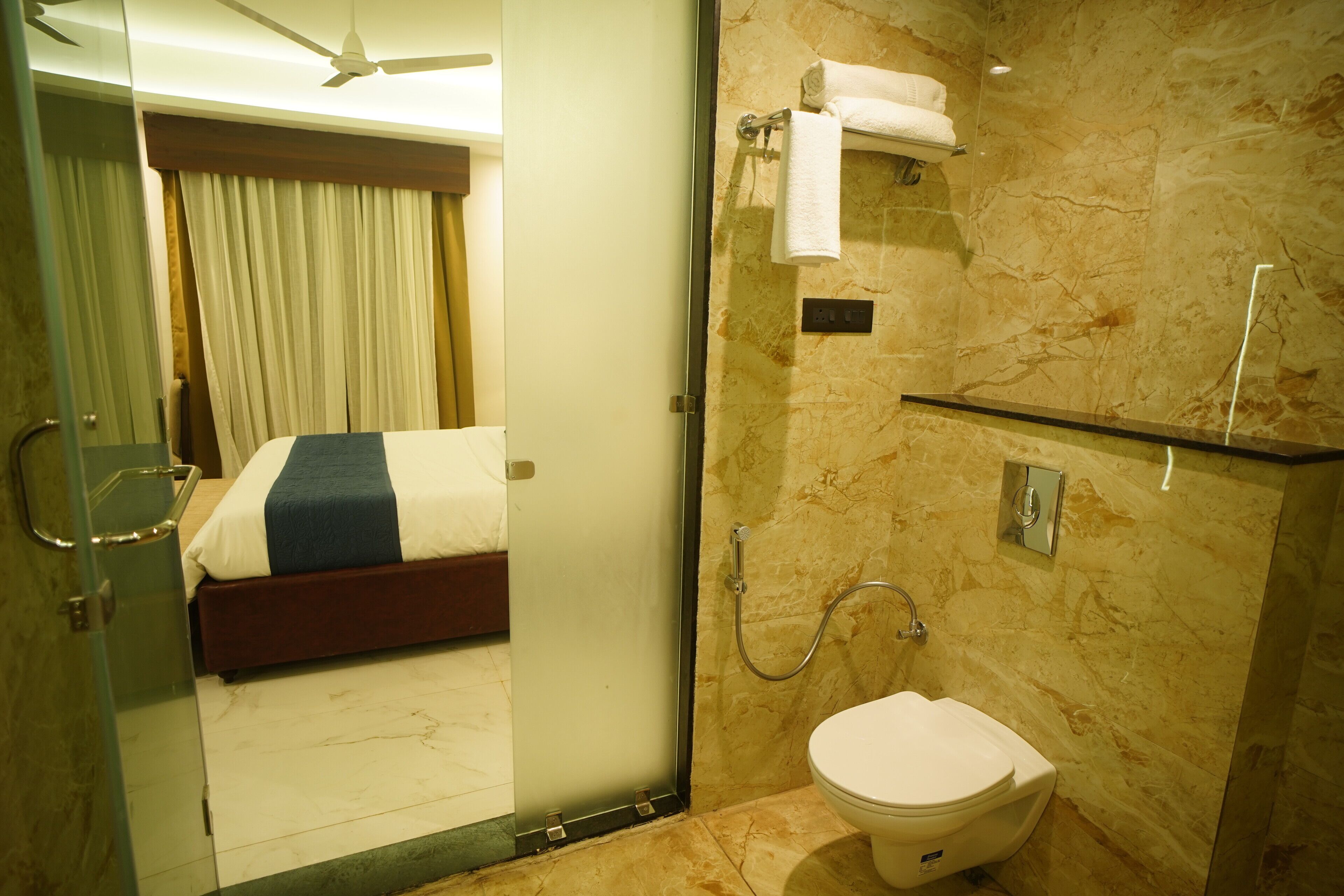 A hotel room with a bed, a glass shower enclosure, a toilet, and a marble wall.