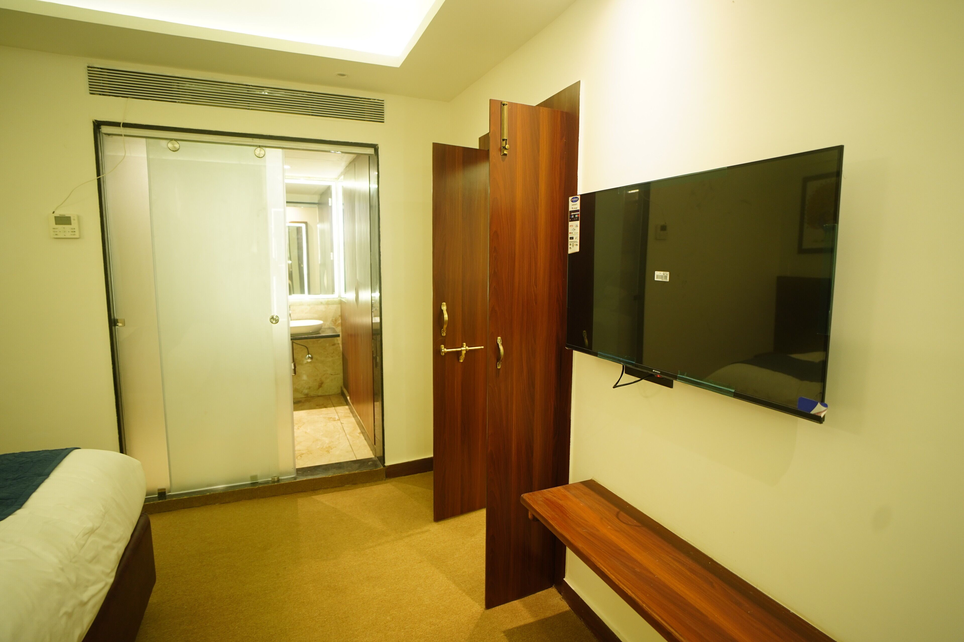 Habitación de hotel con cama, un banco de madera, televisor montado en la pared y una puerta que da al baño.