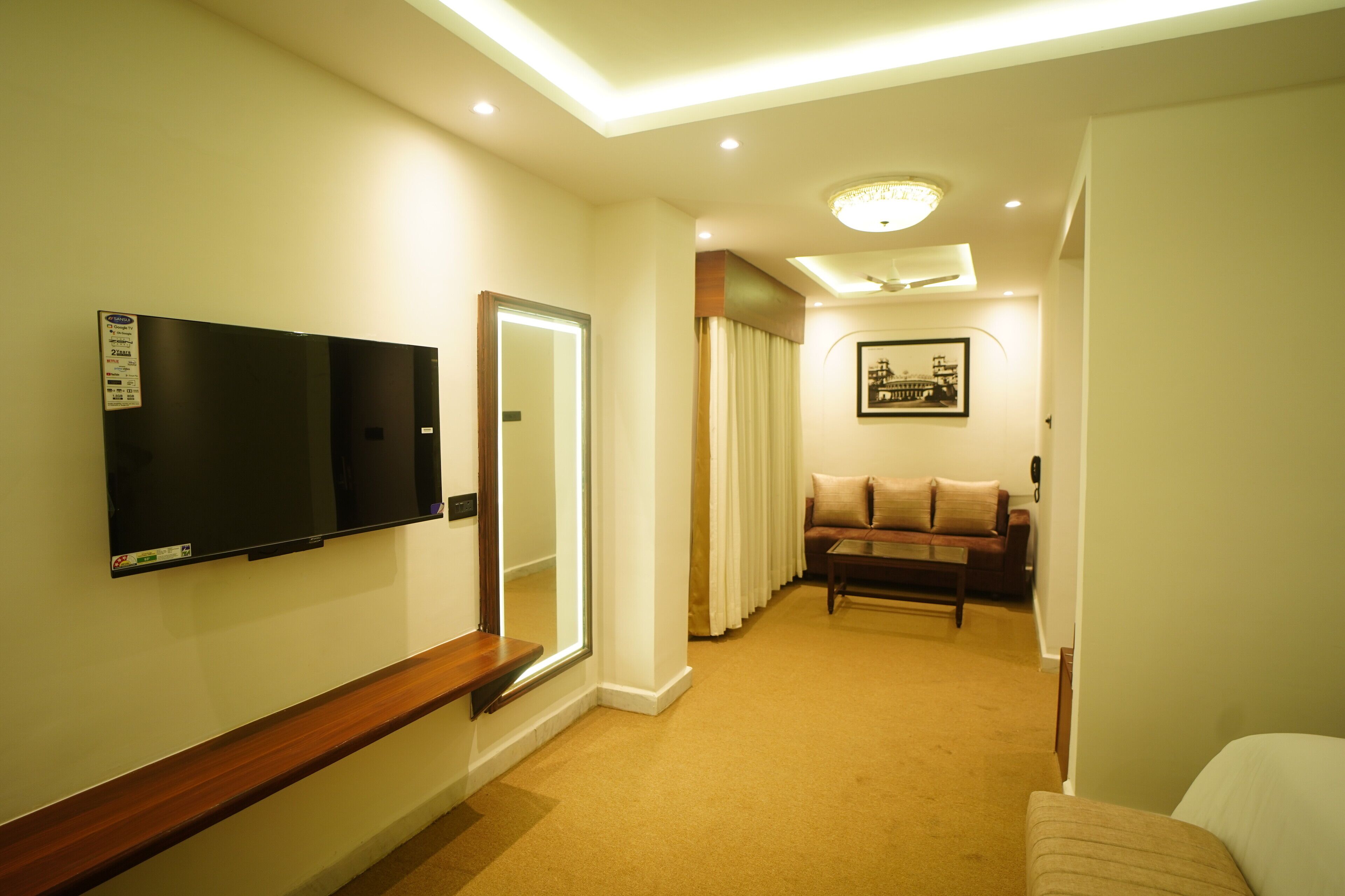 A hotel room with a flat-screen TV mounted on the wall, a large mirror, a sofa, and a framed picture on the wall.