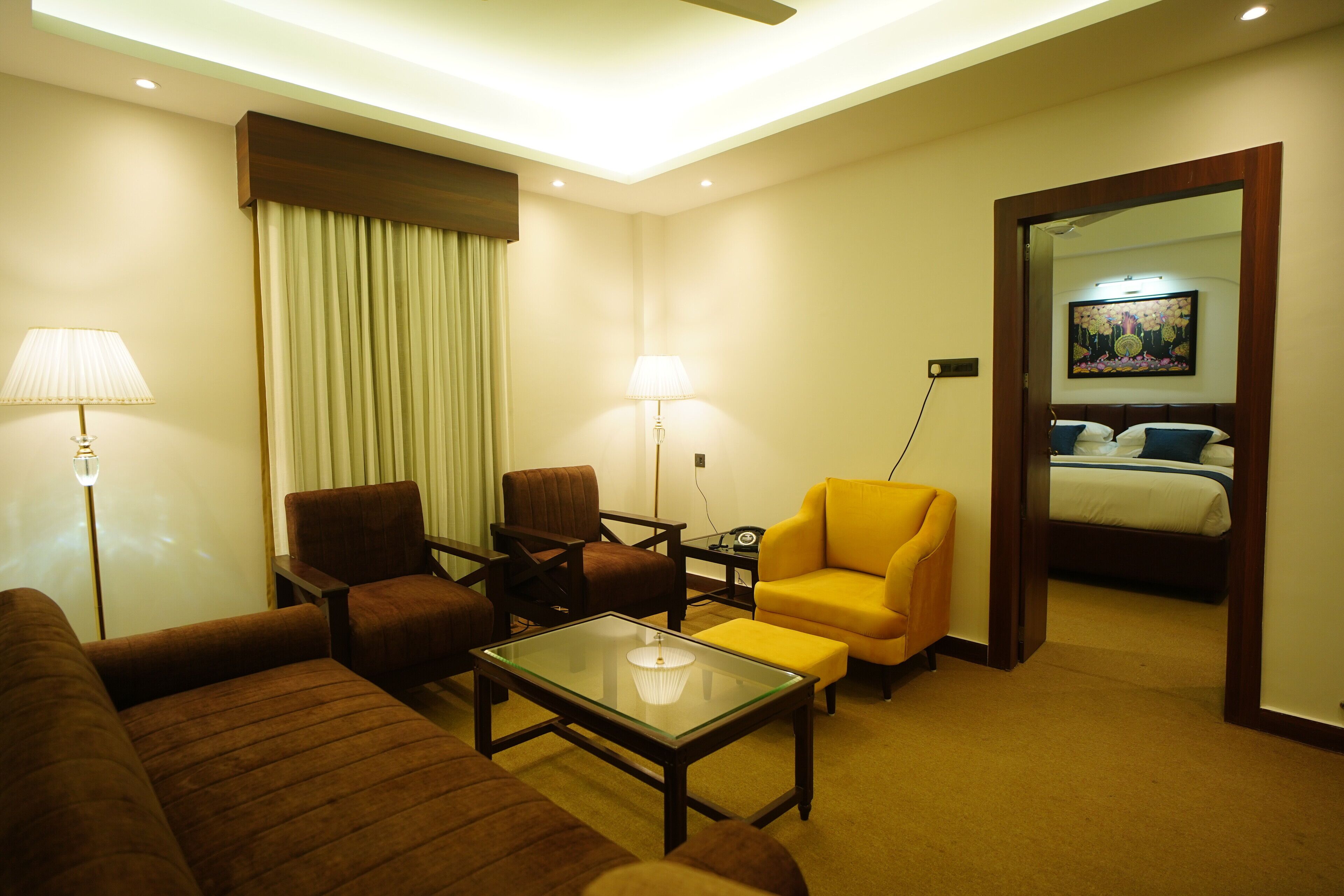 A hotel room with a sofa, armchairs, a coffee table, a lamp, a bed, and a painting on the wall.