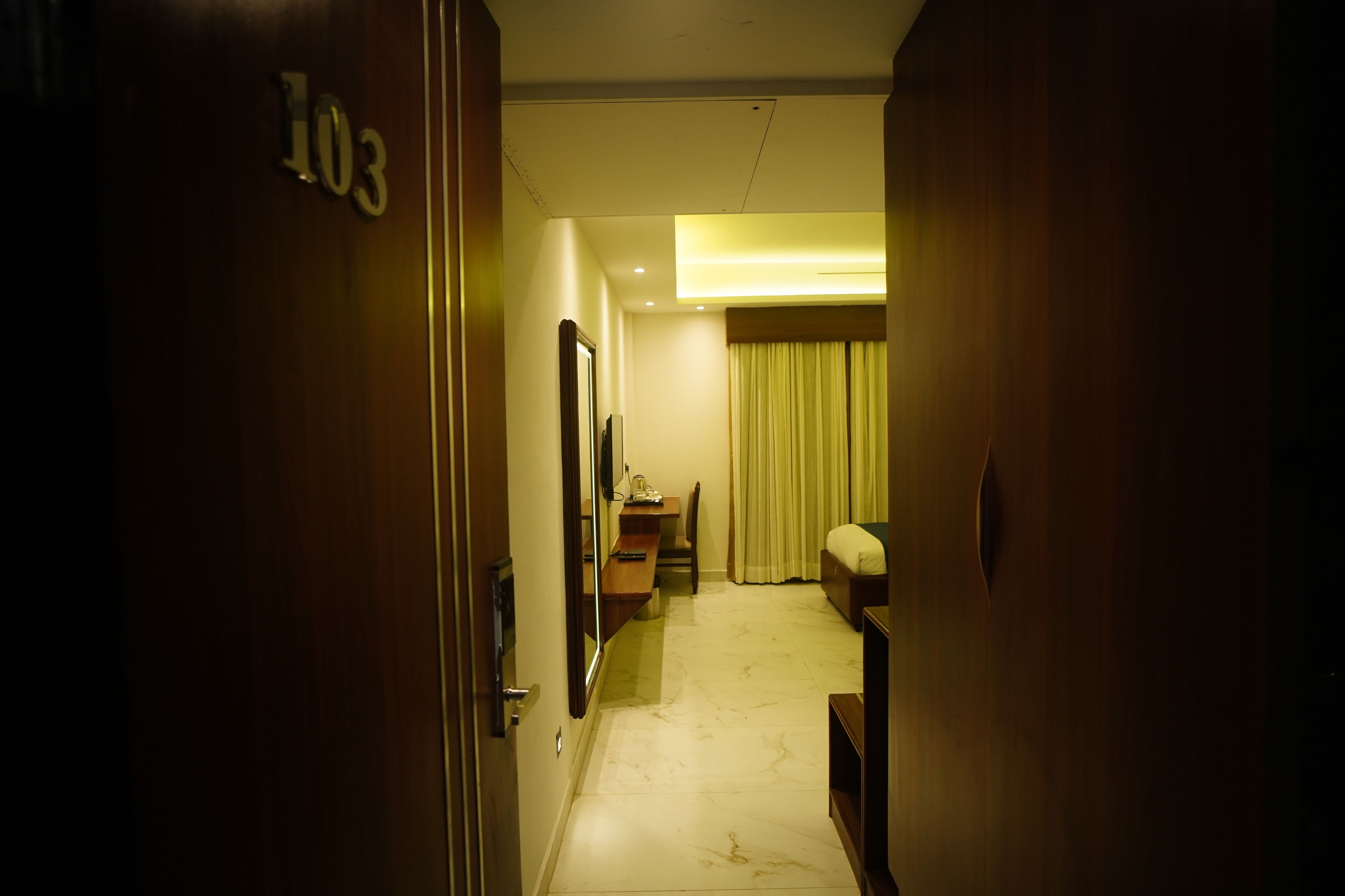 A hotel hallway with a wooden door labeled "103", a desk, a chair, a bed, and a window with curtains.