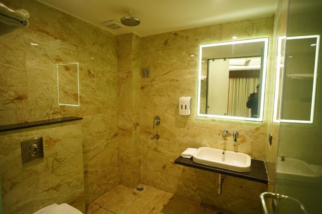 A modern bathroom with a marble wall, a built-in sink, a handheld showerhead, and a mirror with a light.