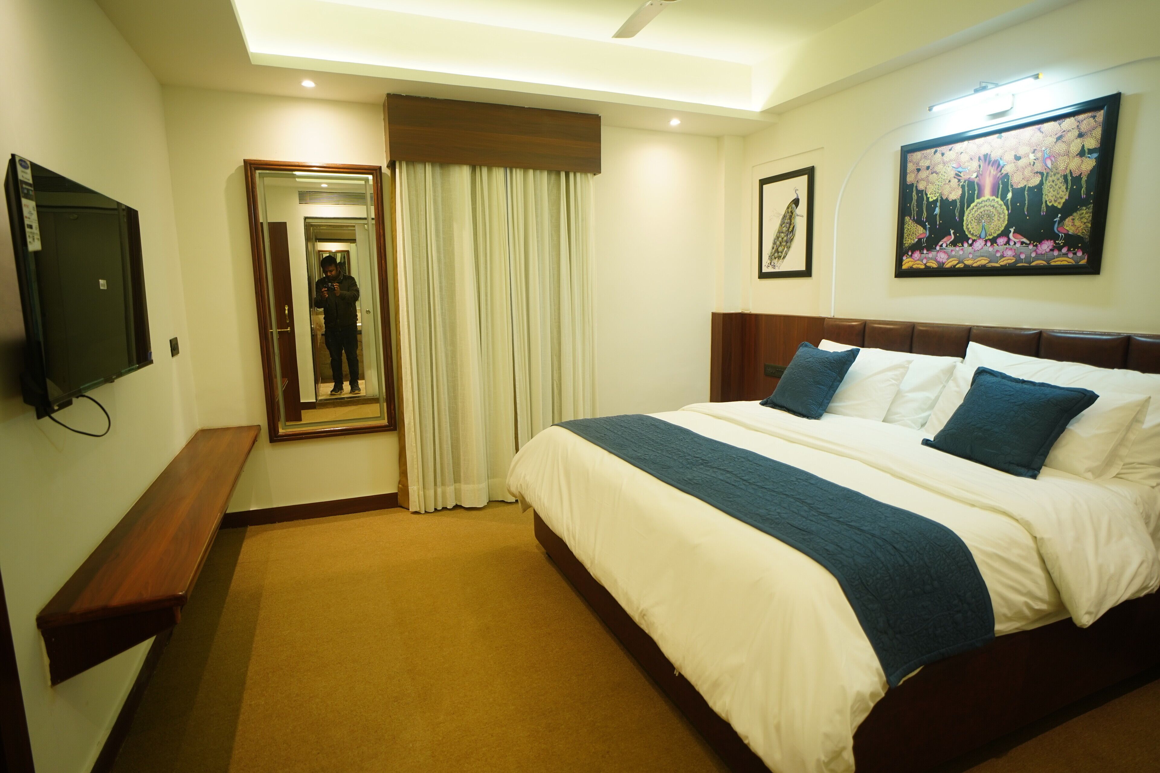 A hotel room with a large bed, a flat-screen TV, a wooden bench, a mirror, and a painting on the wall.